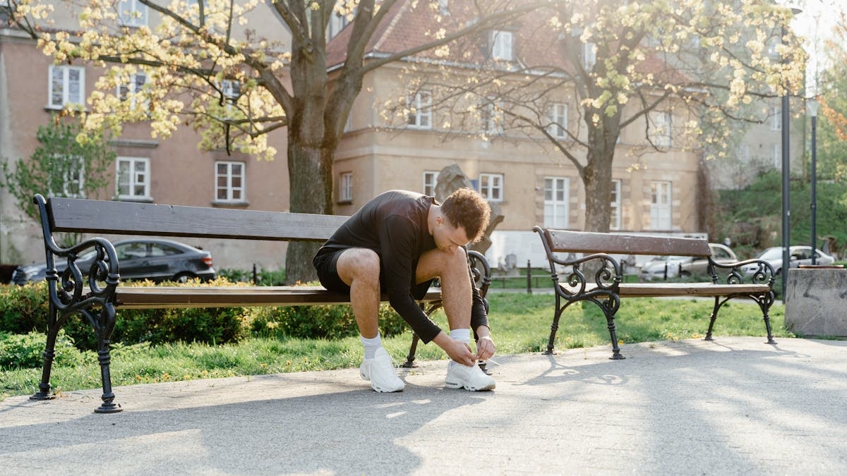 Adult man tying shoes on park bench in morning sunlight, ready for jogging.